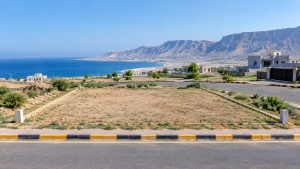 Vacant one-kanal residential plot with concrete boundary markers beside a paved road in Koh-e-Batil, Gwadar, with modern houses, the Arabian Sea, and rugged coastal hills in the background.